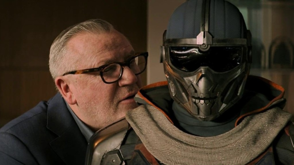 A man stands over the shoulder of a masked creature.