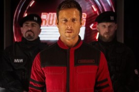 Glen Powell stands in front of two guards.