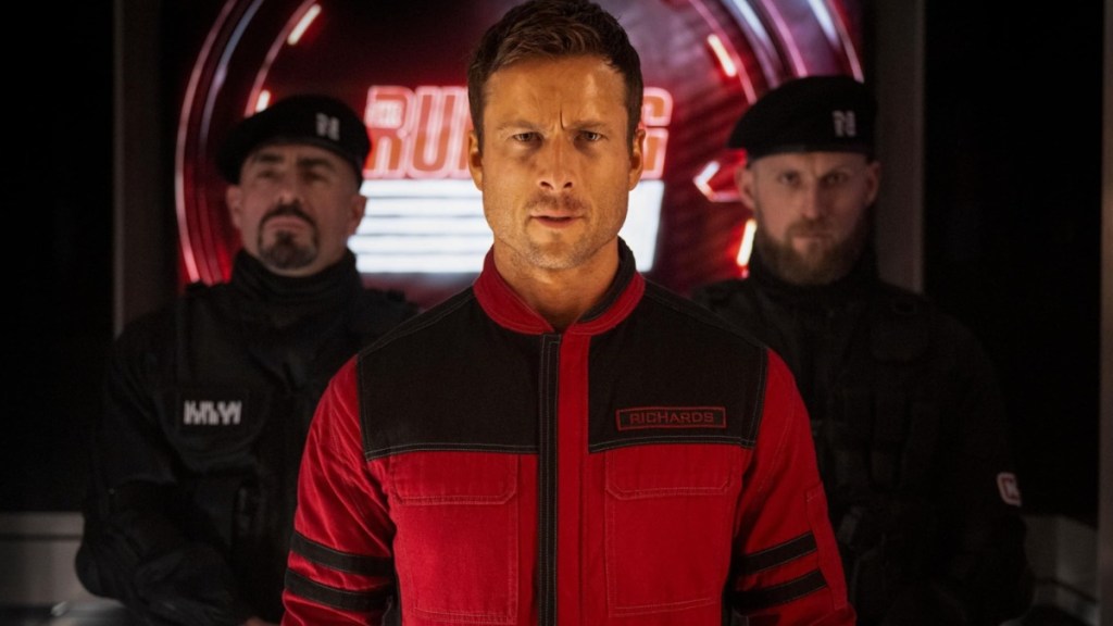 Glen Powell stands in front of two guards.